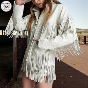 Può includere: Giacca in pelle bianco sporco con lunghe frange sulle maniche, sul davanti e sull'orlo. La giacca ha un collo e tasche. Il logo West Crush è visibile nell'angolo in alto a sinistra. La giacca fa parte di una collezione di abbigliamento di ispirazione western.