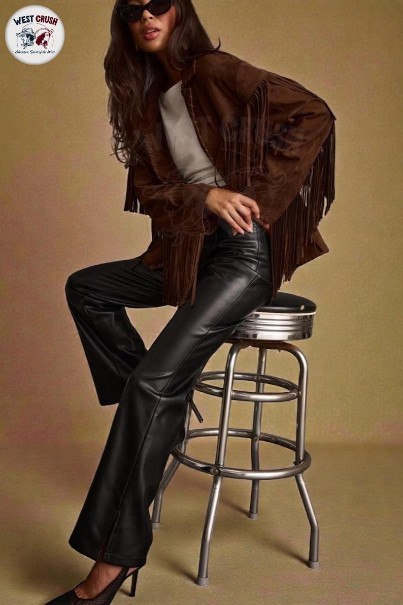 May include: A brown suede jacket featuring long fringe detailing on the sleeves and chest, worn over a plain white top. The outfit includes black leather trousers with a straight-leg cut and black high-heeled shoes. A logo in the corner reads WEST CRUSH.