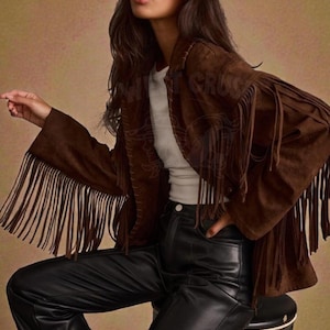 Puede incluir: Chaqueta de ante marrón con flecos largos en las mangas. La chaqueta se combina con pantalones de cuero negros. La modelo lleva gafas de sol negras. El logotipo "West Crush" está en la esquina superior izquierda.