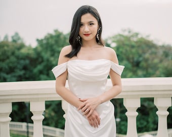 Op de afbeelding: Een witte off-the-shoulder satijnen jurk. De jurk heeft een gedrapeerd lijfje en een zwierige rok. De persoon draagt ​​hangende oorbellen en een ring. De achtergrond omvat een witte balustrade en groen gebladerte.