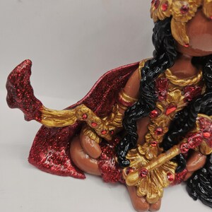 May include: A detailed figurine of a deity, featuring a red and gold cape, ornate gold jewelry with red accents, and black hair. The figure is in a seated position, with intricate detailing on the clothing and accessories.