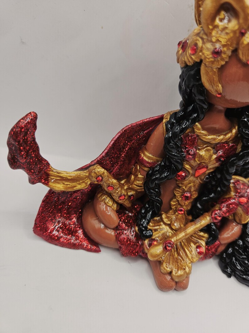 May include: A detailed figurine of a deity, featuring a red and gold cape, ornate gold jewelry with red accents, and black hair. The figure is in a seated position, with intricate detailing on the clothing and accessories.