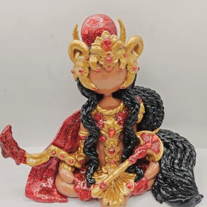May include: A figurine of a seated deity with dark skin, adorned in gold and red. The figure wears a gold crown with red jewels, a red cape, and holds a gold scepter. Long black hair cascades down the back.
