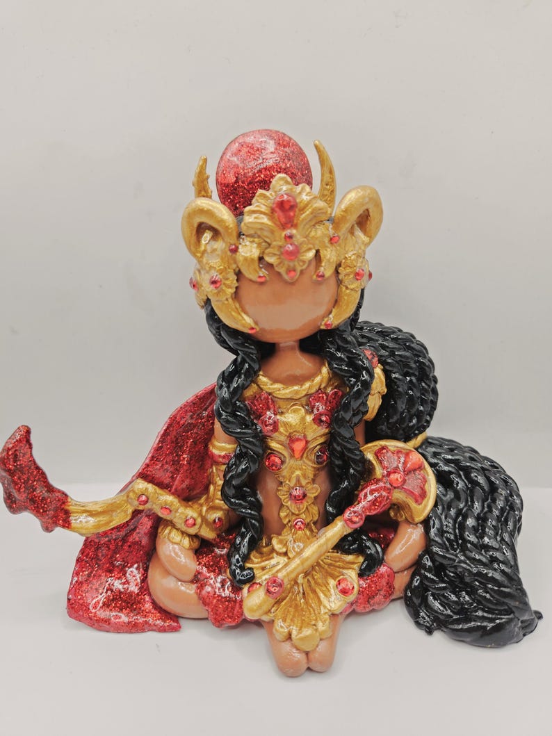 May include: A figurine of a seated deity with dark skin, adorned in gold and red. The figure wears a gold crown with red jewels, a red cape, and holds a gold scepter. Long black hair cascades down the back.