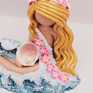 May include: A handcrafted figurine of a mermaid with long, golden hair and a floral crown. The mermaid is holding a seashell with a pearl, and is seated on a wave-like base with blue and white accents. Pink and yellow flowers adorn her dress.