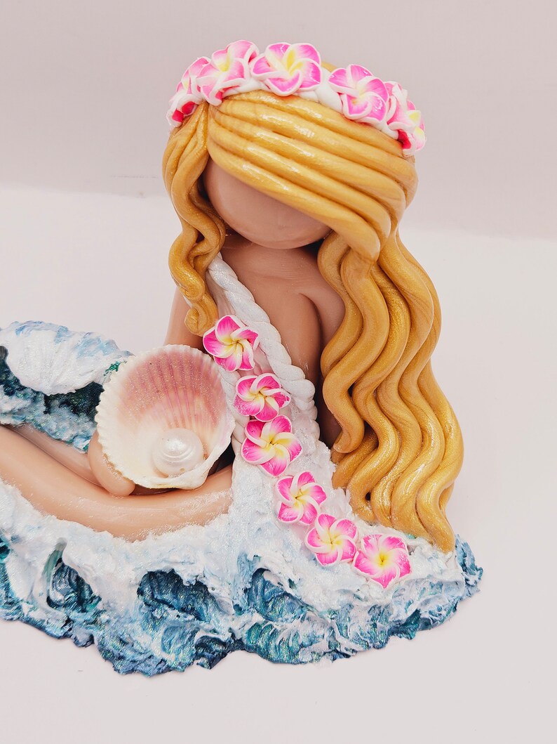 May include: A handcrafted figurine of a mermaid with long, golden hair and a floral crown. The mermaid is holding a seashell with a pearl, and is seated on a wave-like base with blue and white accents. Pink and yellow flowers adorn her dress.
