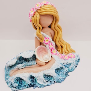 May include: A handcrafted figurine of a mermaid with long blonde hair, a floral crown, and a seashell. The mermaid is seated on a sculpted wave with blue and white accents, holding a seashell with a pearl.
