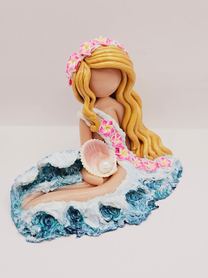 May include: A handcrafted figurine of a mermaid with long blonde hair, a floral crown, and a seashell. The mermaid is seated on a sculpted wave with blue and white accents, holding a seashell with a pearl.