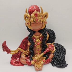 May include: A handcrafted figurine of a deity, adorned in gold and red. The figure wears a jeweled crown and cape, with long black hair. The figurine is seated, holding a staff, and has a red orb atop its head.