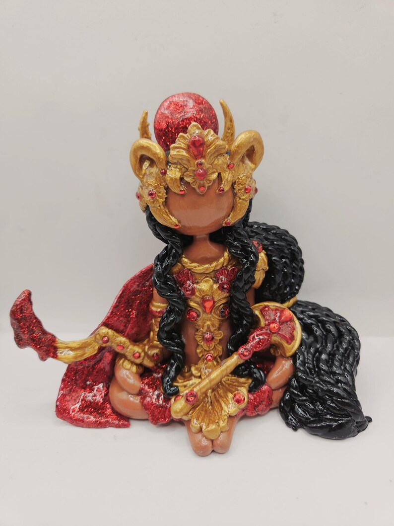 May include: A handcrafted figurine of a deity, adorned in gold and red. The figure wears a jeweled crown and cape, with long black hair. The figurine is seated, holding a staff, and has a red orb atop its head.
