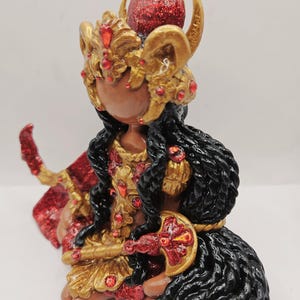 May include: A handcrafted figurine of a seated figure with dark skin, adorned in gold and red accents. The figure wears a detailed headdress with horns and a red glitter sphere. The figure's hair is black and wavy, and it holds a decorative object.