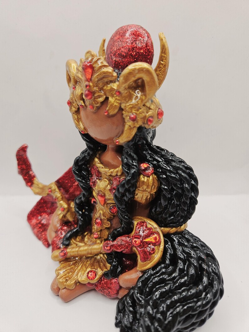 May include: A handcrafted figurine of a seated figure with dark skin, adorned in gold and red accents. The figure wears a detailed headdress with horns and a red glitter sphere. The figure's hair is black and wavy, and it holds a decorative object.