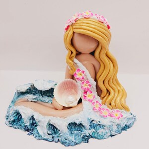 May include: A handcrafted figurine of a mermaid with long blonde hair, a floral crown, and a seashell. The mermaid is seated on a blue and white wave base, holding a pink seashell. The figurine is a decorative item.