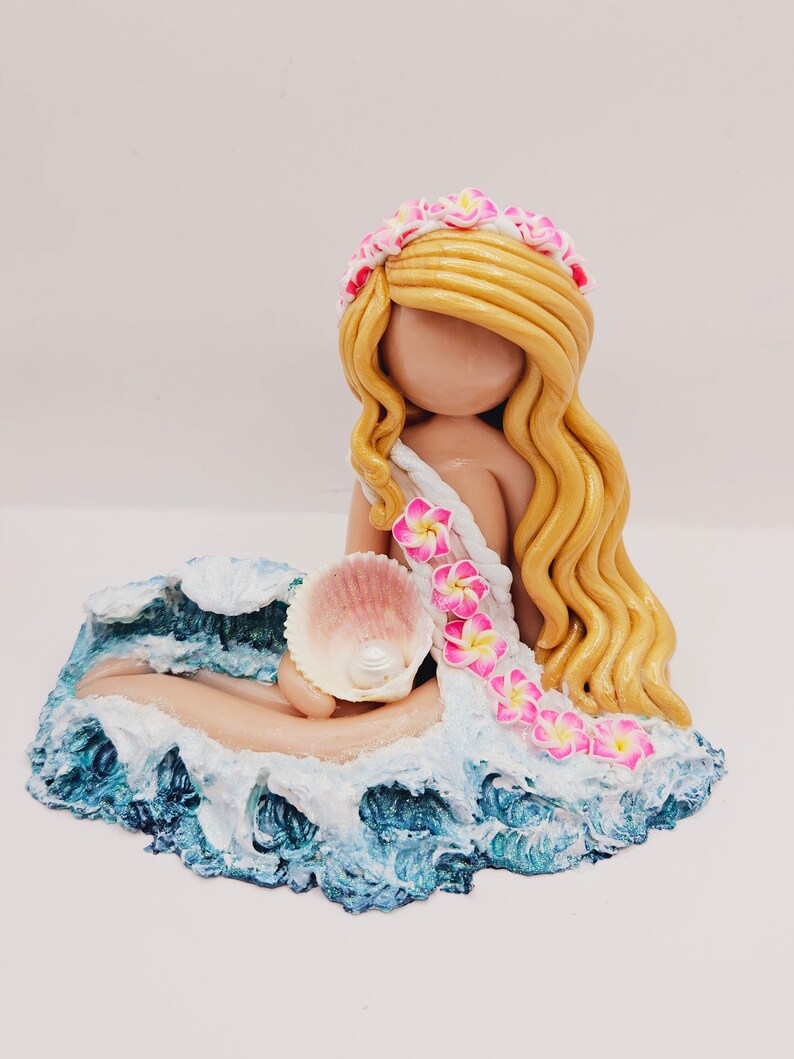 May include: A handcrafted figurine of a mermaid with long blonde hair, a floral crown, and a seashell. The mermaid is seated on a blue and white wave base, holding a pink seashell. The figurine is a decorative item.