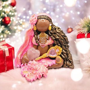 May include: A handcrafted figurine of a seated woman with brown skin, wearing a pink head wrap, dress, and holding a heart-shaped stone and a golden mirror. The figurine has long, braided brown hair and gold earrings. The background includes a Christmas tree and wrapped gifts.