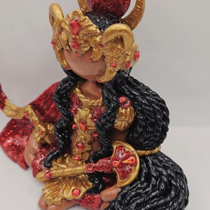 May include: A detailed figurine of a deity, featuring a dark complexion and adorned with gold and red accents. The figure wears a gold headdress with red jewels, a red cape, and holds a gold and red weapon. The hair is black and the figure is seated.