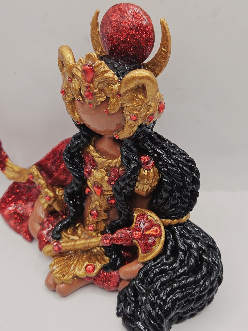 May include: A detailed figurine of a deity, featuring a dark complexion and adorned with gold and red accents. The figure wears a gold headdress with red jewels, a red cape, and holds a gold and red weapon. The hair is black and the figure is seated.