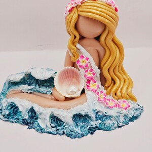 May include: A handcrafted figurine of a mermaid with long, flowing blonde hair and a floral crown. She holds a seashell with a pearl, resting on a base of ocean waves. The mermaid's dress is adorned with pink and white flowers.
