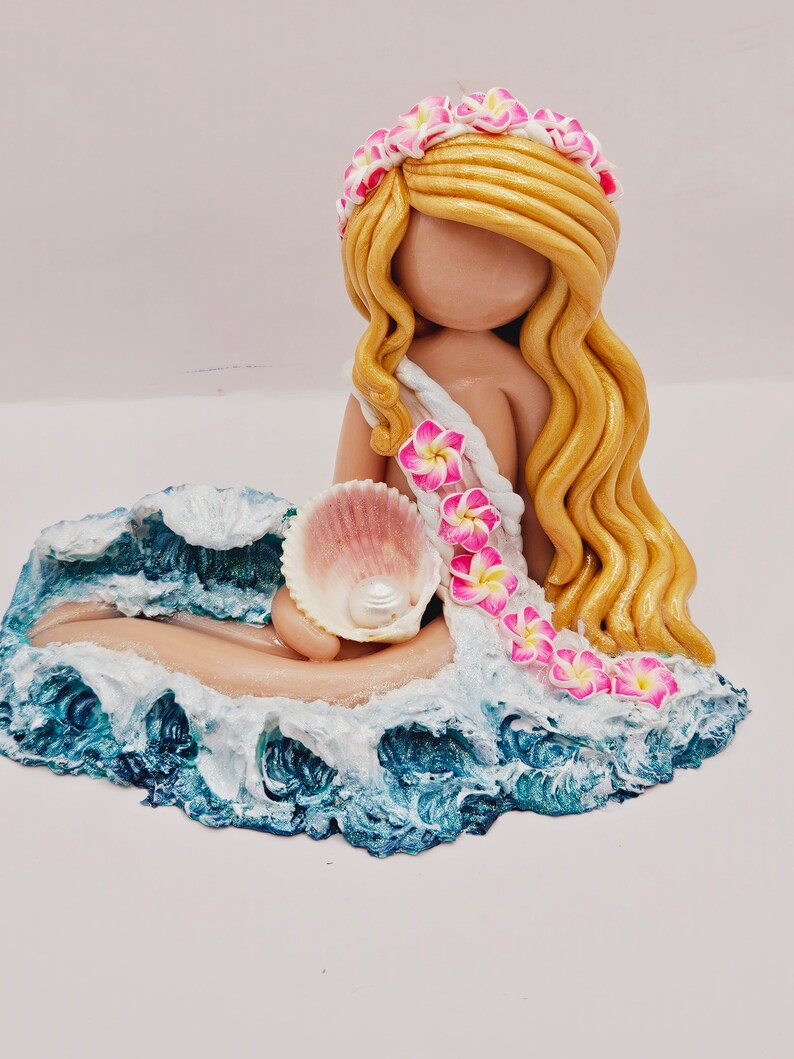 May include: A handcrafted figurine of a mermaid with long, flowing blonde hair and a floral crown. She holds a seashell with a pearl, resting on a base of ocean waves. The mermaid's dress is adorned with pink and white flowers.