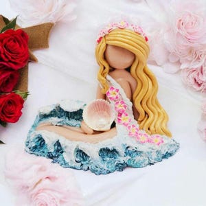 May include: A handcrafted mermaid figurine with long blonde hair and a floral crown. The mermaid is seated on a blue and white wave base, holding a seashell. The figurine's dress is adorned with pink flowers.