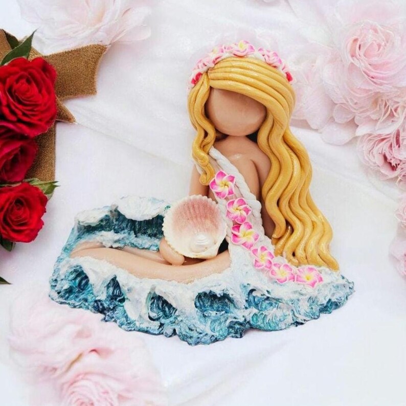 May include: A handcrafted mermaid figurine with long blonde hair and a floral crown. The mermaid is seated on a blue and white wave base, holding a seashell. The figurine's dress is adorned with pink flowers.