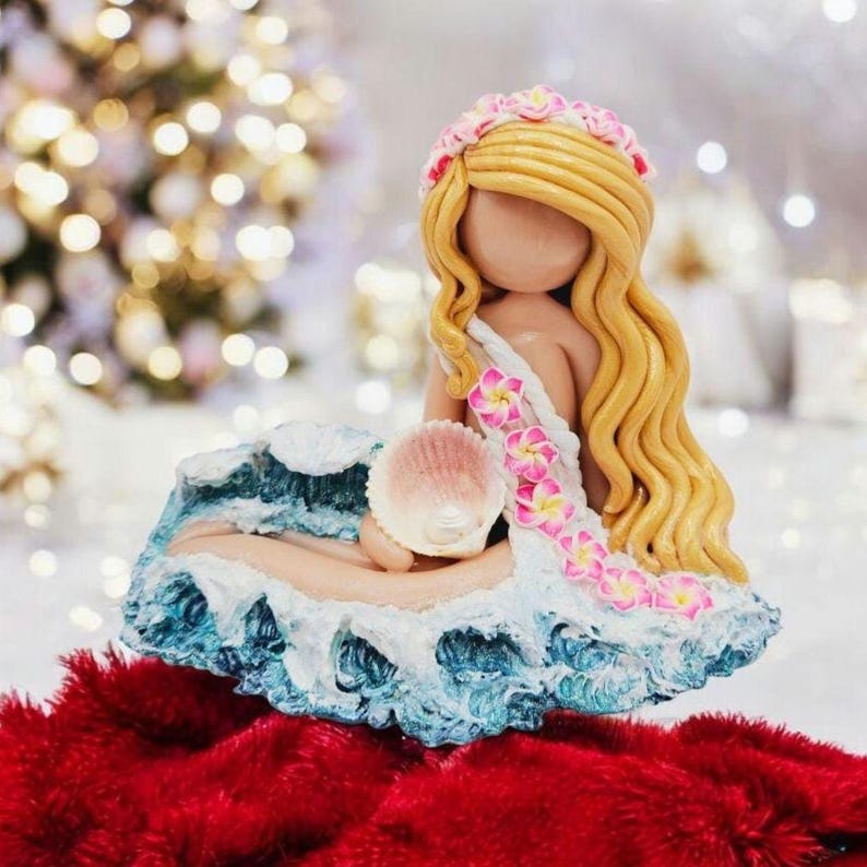 May include: A handcrafted figurine of a mermaid with long blonde hair, a floral crown, and a seashell. The mermaid is seated on a wave-like base with blue and white details, and is wearing a floral garland.