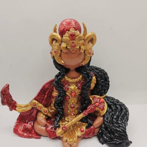 May include: A handcrafted figurine of a seated deity, adorned in gold and red. The figure wears a jeweled crown and cape, with long black hair. The figurine holds a staff and has a red orb on its head.