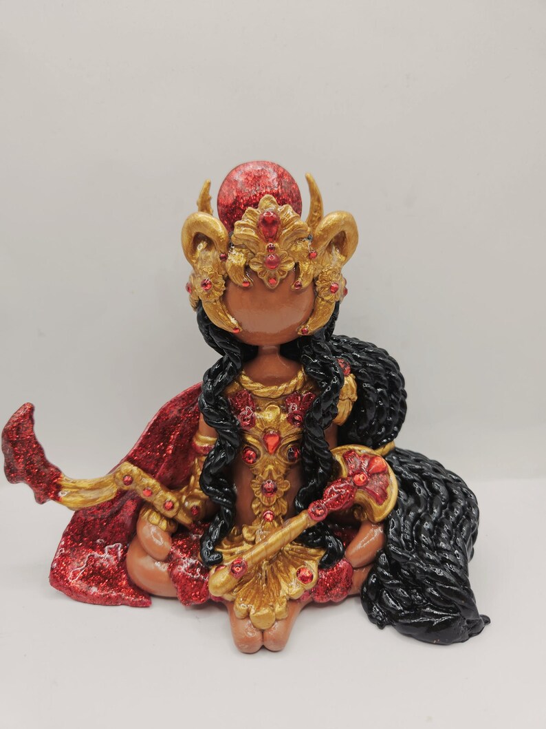 May include: A handcrafted figurine of a seated deity, adorned in gold and red. The figure wears a jeweled crown and cape, with long black hair. The figurine holds a staff and has a red orb on its head.
