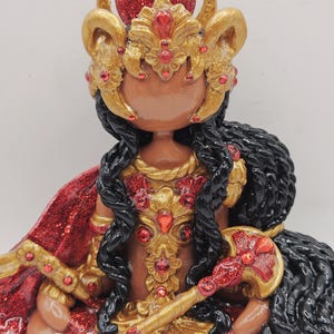 May include: A handcrafted figurine of a deity, featuring a brown face, black braided hair, and elaborate gold and red accents. The figure wears a gold crown with red jewels and a red glitter cape. The figurine is seated.