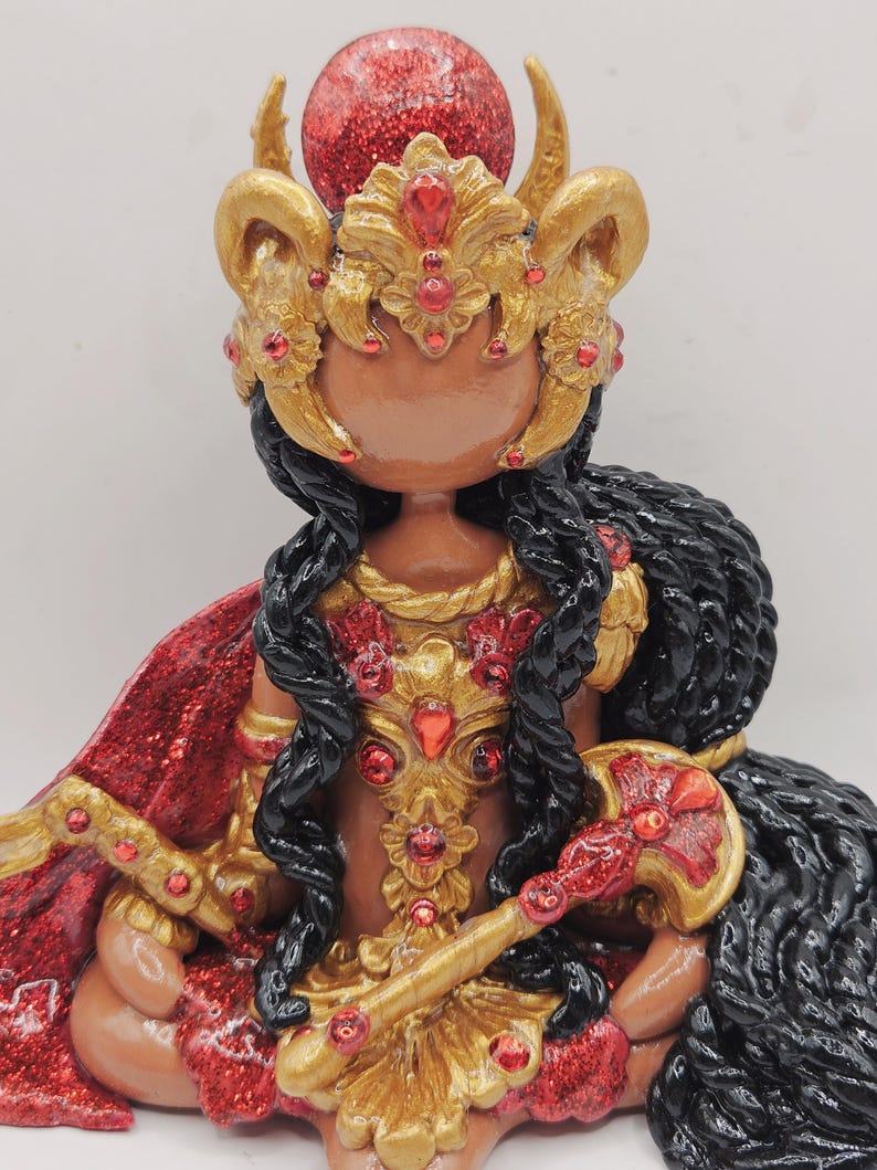 May include: A handcrafted figurine of a deity, featuring a brown face, black braided hair, and elaborate gold and red accents. The figure wears a gold crown with red jewels and a red glitter cape. The figurine is seated.