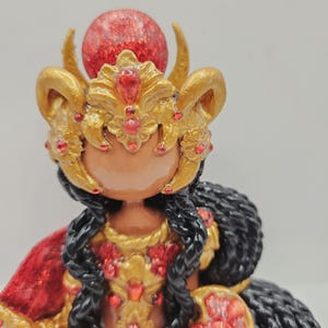 May include: A detailed figurine of a deity, featuring a gold headdress with red accents and a large red orb. The figure has black hair and wears a red and gold garment. The overall design is ornate and decorative.
