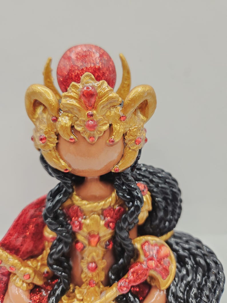 May include: A detailed figurine of a deity, featuring a gold headdress with red accents and a large red orb. The figure has black hair and wears a red and gold garment. The overall design is ornate and decorative.
