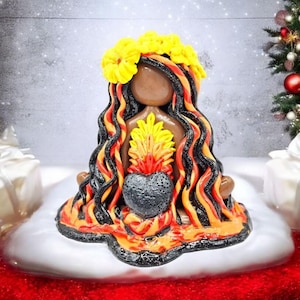 Puede incluir: Figura artesanal de una mujer con largo cabello ondulado negro, naranja y amarillo, con una corona de flores amarillas. Sostiene un objeto en forma de corazón con un diseño de llama. La figura está sobre una superficie blanca.