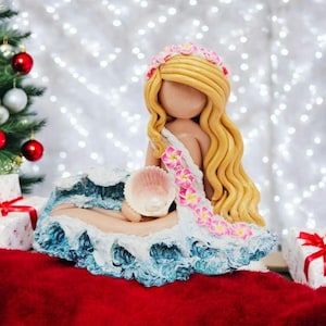 May include: A handcrafted mermaid figurine with long blonde hair, a floral crown, and a seashell. The mermaid is seated on a wave-like base with blue and white details. The figurine is decorated with pink flowers.