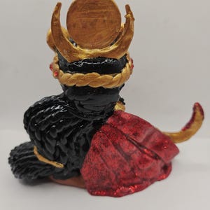 May include: A handcrafted figurine of a seated figure with black hair and a red cape. The figure wears a gold crown with a circular emblem and a gold braided headpiece. Red gems are set into the headpiece.