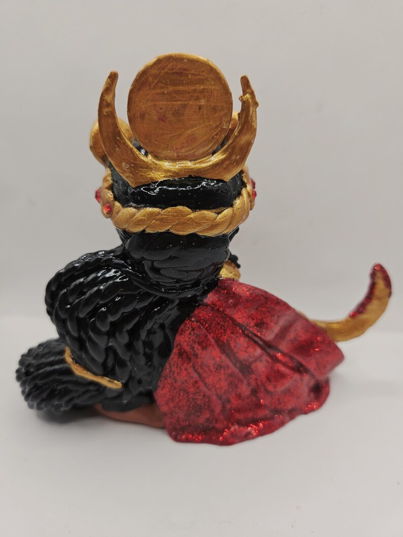May include: A handcrafted figurine of a seated figure with black hair and a red cape. The figure wears a gold crown with a circular emblem and a gold braided headpiece. Red gems are set into the headpiece.