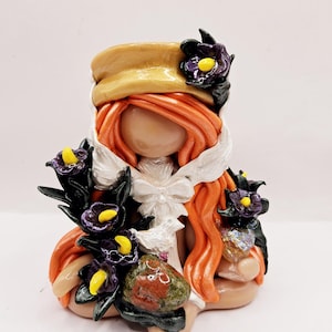 May include: A handmade figurine of a seated figure with orange hair, wearing a tan hat and white collar. The figurine is decorated with purple and yellow flowers and holds a decorative stone. The overall theme is whimsical.