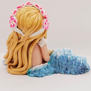 May include: A handcrafted figurine of a mermaid with long, flowing blonde hair and a floral crown. The mermaid is seated on a blue and white wave, with her back to the viewer. The figurine is made of a smooth, glossy material.