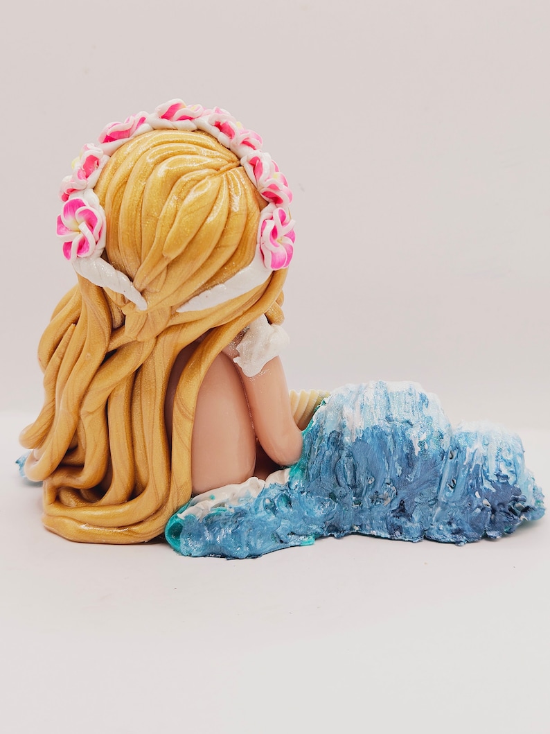 May include: A handcrafted figurine of a mermaid with long, flowing blonde hair and a floral crown. The mermaid is seated on a blue and white wave, with her back to the viewer. The figurine is made of a smooth, glossy material.