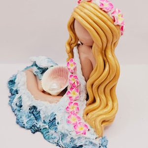 May include: A handcrafted figurine of a mermaid with long, golden hair and a floral crown. The mermaid is nestled in a sculpted wave with blue and white hues, holding a seashell. Pink and yellow flowers adorn the mermaid's dress.