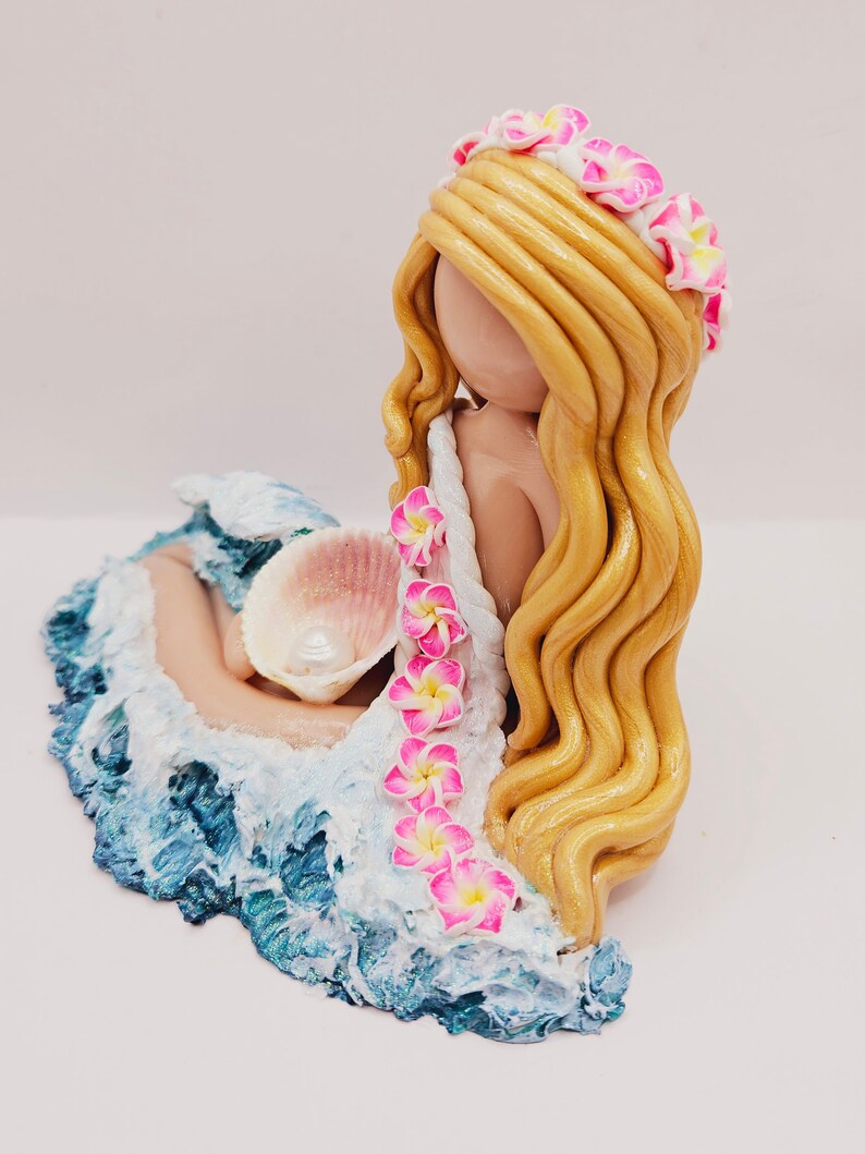 May include: A handcrafted figurine of a mermaid with long, golden hair and a floral crown. The mermaid is nestled in a sculpted wave with blue and white hues, holding a seashell. Pink and yellow flowers adorn the mermaid's dress.