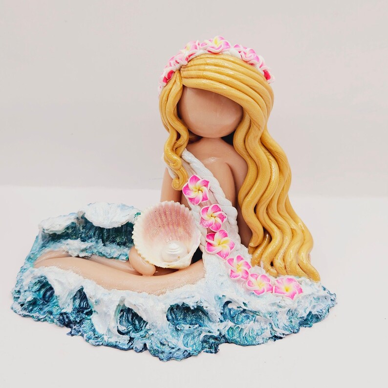May include: A handcrafted figurine of a mermaid with long blonde hair and a floral crown. She holds a seashell and is seated on a wave-like base with blue and white accents. The mermaid's attire is adorned with pink and yellow flowers.