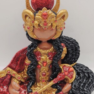May include: A detailed figurine of a deity, featuring a red glittery orb atop a golden headdress with curved horns. The figure has black hair, a red and gold cape, and holds a golden scepter. Red gemstones adorn the golden armor and headdress.