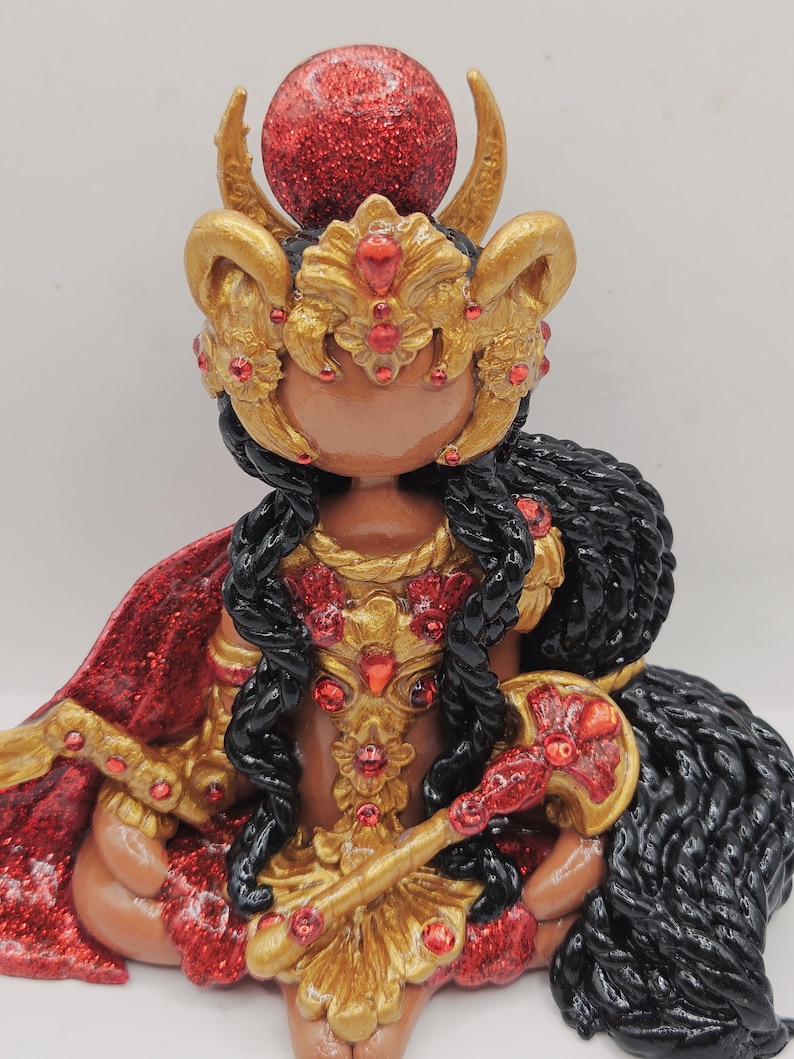 May include: A detailed figurine of a deity, featuring a red glittery orb atop a golden headdress with curved horns. The figure has black hair, a red and gold cape, and holds a golden scepter. Red gemstones adorn the golden armor and headdress.