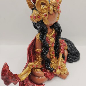 May include: A handcrafted figurine of a seated figure with dark skin, adorned in gold and red attire. The figure wears a detailed headdress and cape, with long black braids. The figurine is embellished with red glitter and jewels.