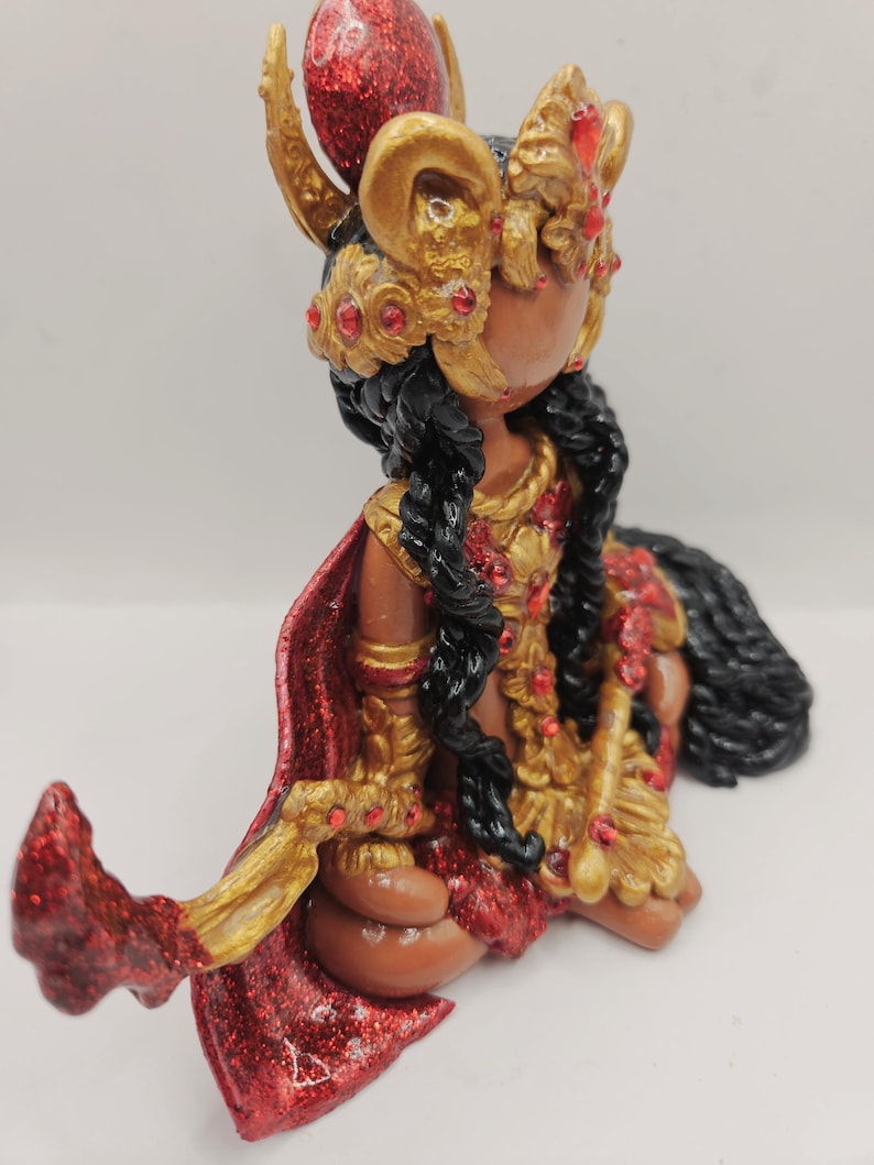May include: A handcrafted figurine of a seated figure with dark skin, adorned in gold and red attire. The figure wears a detailed headdress and cape, with long black braids. The figurine is embellished with red glitter and jewels.