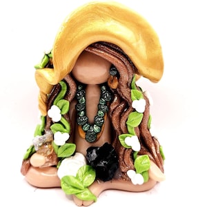 May include: A handmade figurine of a seated woman with long brown hair, wearing a gold hat, green and white floral accents, and a green beaded necklace. The figurine also features a black stone and a white flower.