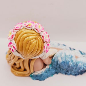 May include: A handcrafted figurine of a person with golden hair, wearing a floral crown of pink and white flowers. The figure is lying down, with a blue and white wave design at the base.