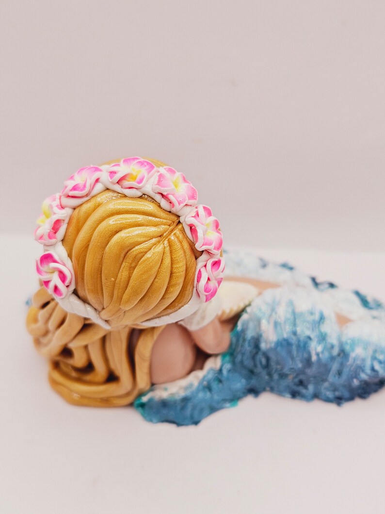 May include: A handcrafted figurine of a person with golden hair, wearing a floral crown of pink and white flowers. The figure is lying down, with a blue and white wave design at the base.