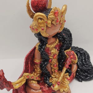 May include: A handcrafted figurine of a seated figure with dark skin, wearing a gold and red ornate headdress and clothing. The figurine has long black hair and a red cape. The figure is holding a gold staff with red accents. The figurine is on a white surface.